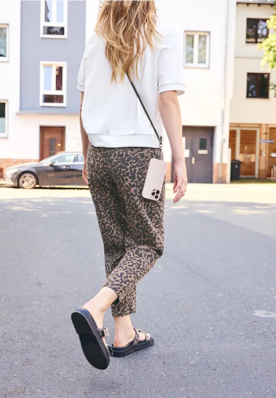 Femme marche dans la rue, porte un pantalon léopard, chemise blanche, sandales noires et tient un téléphone.