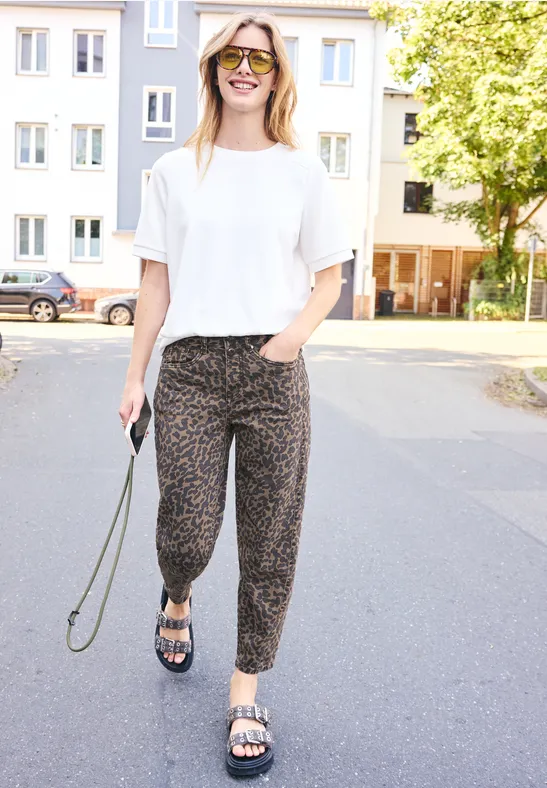 Femme en t-shirt blanc, pantalon à motifs et sandales se promène par une rue résidentielle ensoleillée.