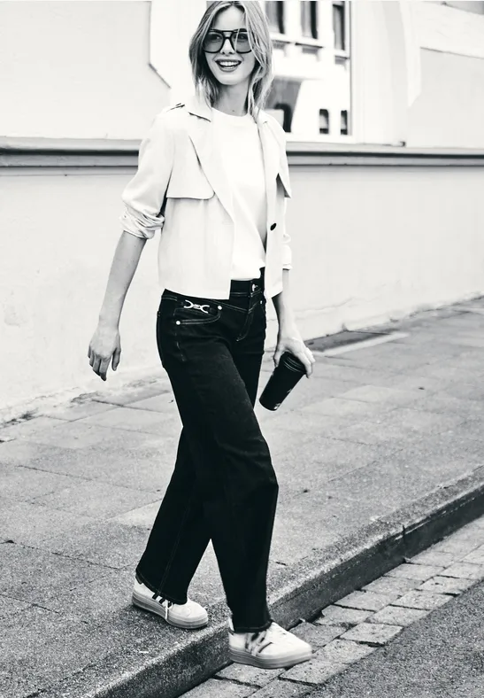 Femme souriante en jeans,veste légère,baskets et lunettes de soleil marche en tenant une pochette.