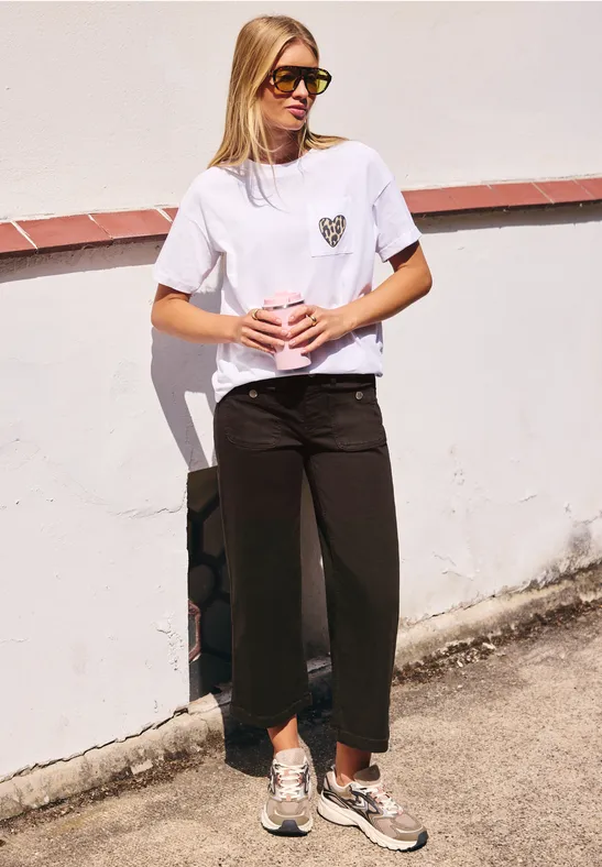 Blonde Frau mit Sonnenbrille, weißem T-Shirt und schwarzen Hosen steht entspannt vor weißer Wand.