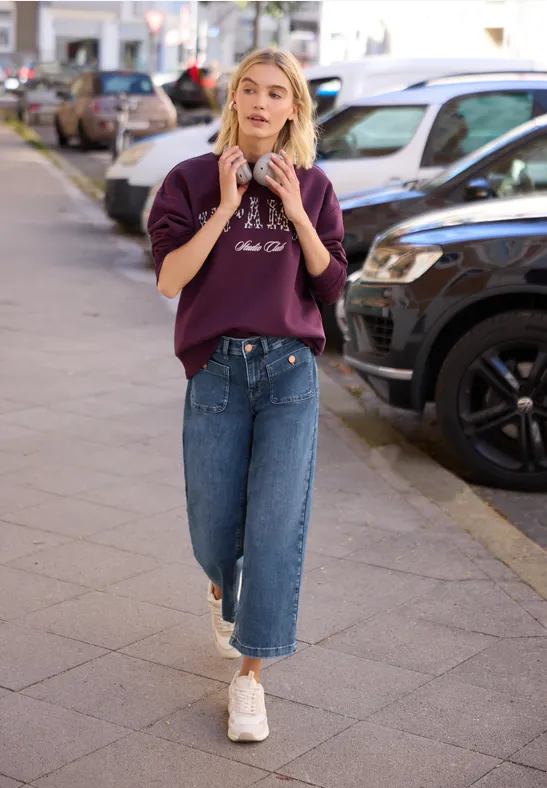 Eine junge Frau in Kapuzenpullover, Jeans und Sneakern läuft auf einem Bürgersteig.