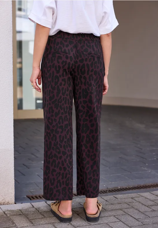 Une personne vue de dos porte un pantalon large à motif léopard rouge et des sandales dans un cadre urbain.