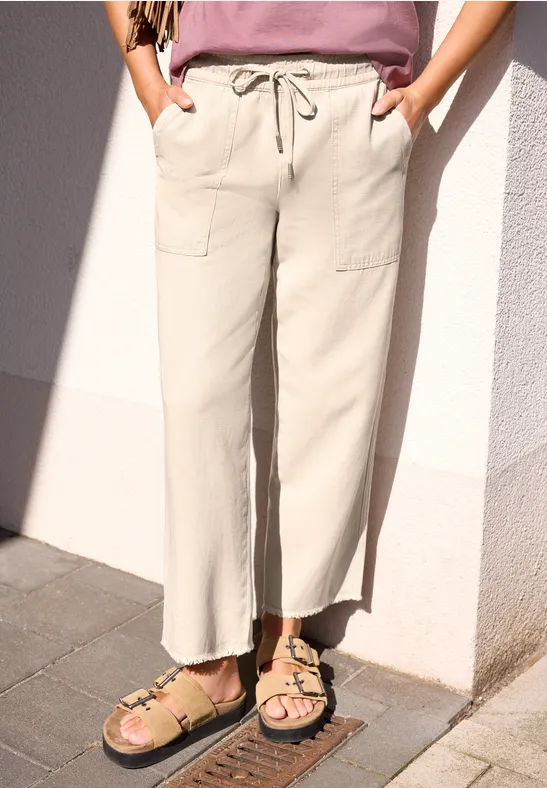 Vrouw in beige, wijde broek met koord en zakken, sandalen aan, voor een witte muur.