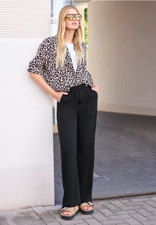Femme appuyée contre un mur dans un couloir, pantalon noir ample, chemisier imprimé et sandales.