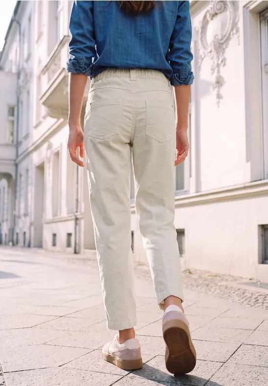 Une personne de dos, en pantalon clair et veste en jean, marche dans une rue ensoleillée.