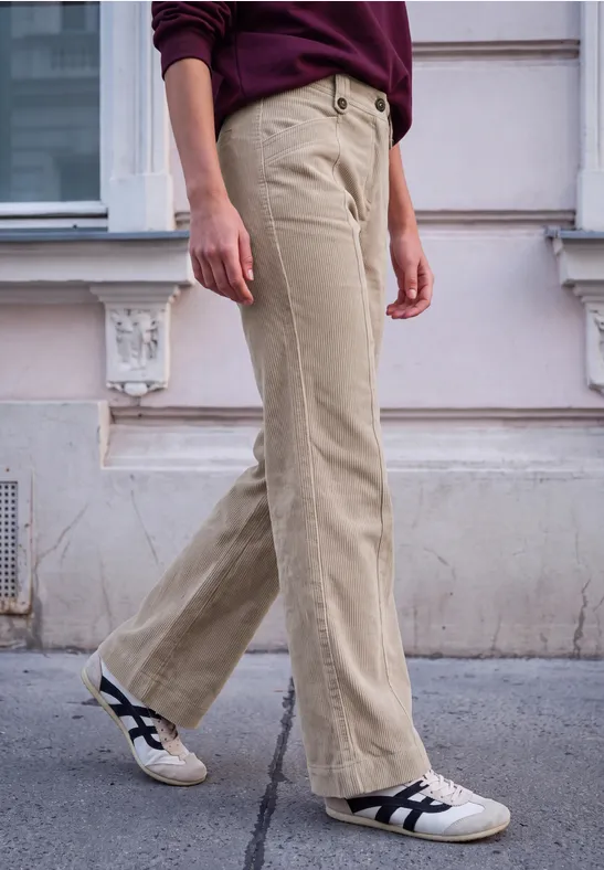 Personne en pantalon velours beige clair et baskets blanches marche devant une façade.