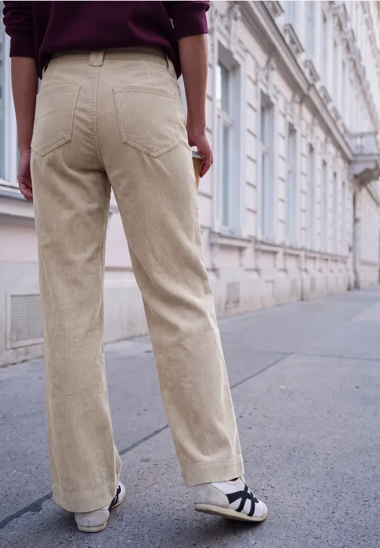 Personne en pantalon en velours beige et baskets pose sur un trottoir devant une façade classique.