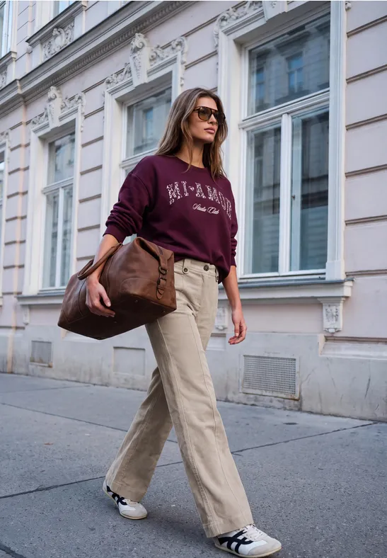 Une femme élégante en pantalon beige, sweat bordeaux et lunettes de soleil marche près d’une façade