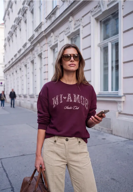 Femme en lunettes de soleil, sweat bordeaux 'Mi AMOR', pantalon beige, sac et téléphone, marchant dans une rue urbaine.