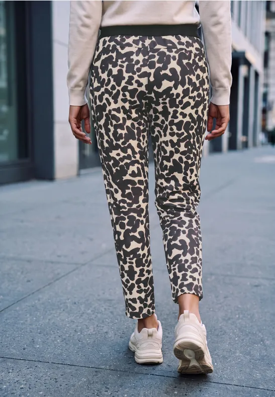 Eine Person geht auf einem Gehweg, trägt enge, gemusterte Hose mit Leopardenflecken und weiße Sneaker.