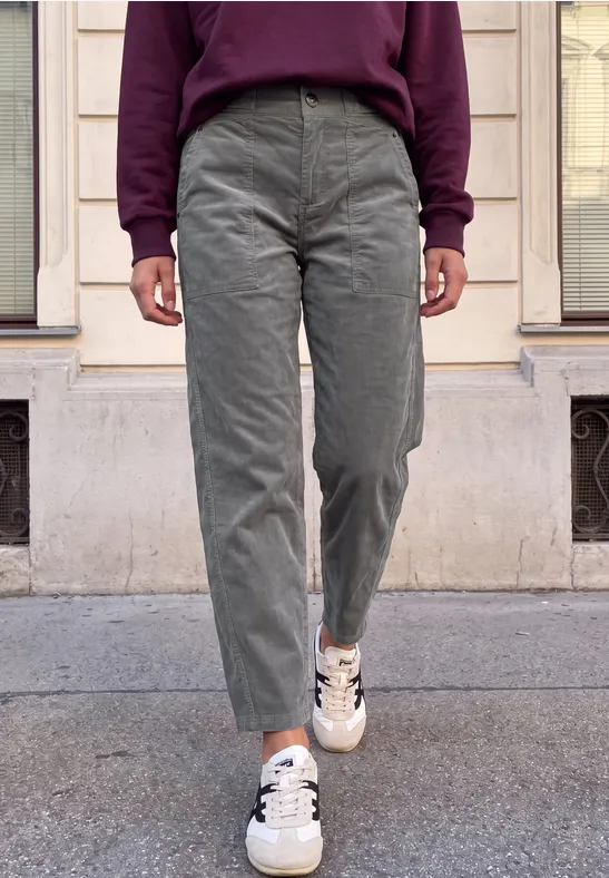 Person in grüner Cordhose, weiß-schwarzen Sneakern und bordeauxfarbenem Pullover steht auf dem Gehweg vor Hausfassade.