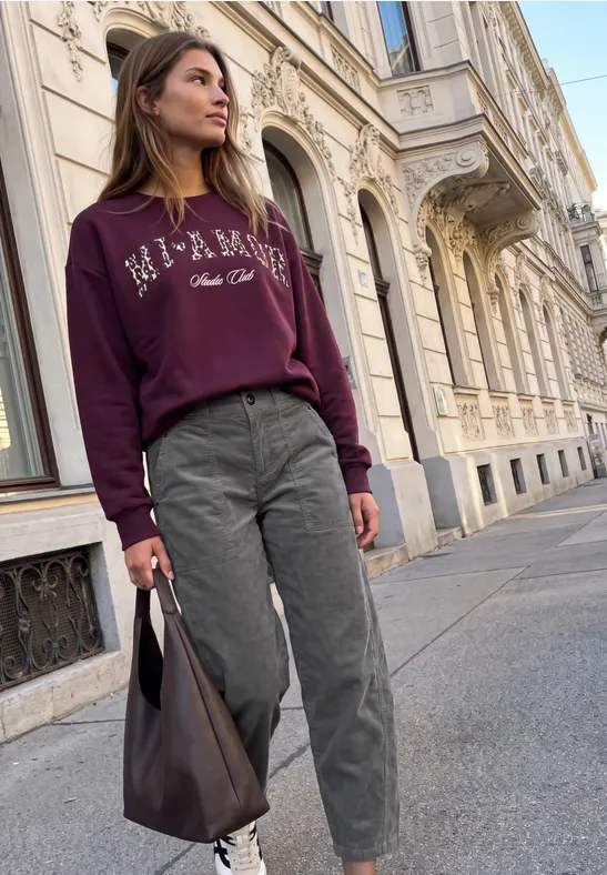 Eine Frau mit burgunderrotem Pullover, grauen Hosen und Ledertasche spaziert vor klassischer Stadtkulisse.