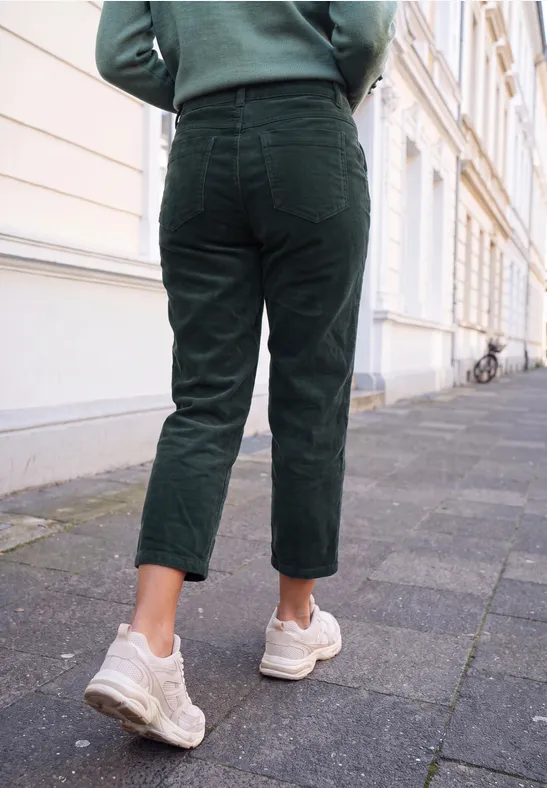 Eine Person in dunkelgrüner Cordhose und weißen Sneakern geht eine Straße neben Häusern entlang.