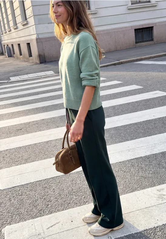 Frau im mintgrünen Pullover, dunklen Hosen und weißen Sneakers, mit brauner Tasche, wartend auf einem Zebrastreifen.