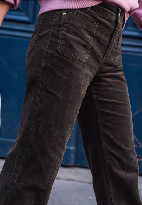 Dunkelbraune Cordhose, getragen von einer Person vor einer blauen Tür, mit pinkem Oberteil erkennbar.