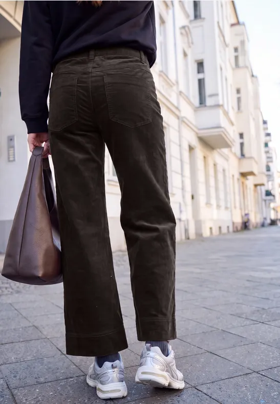 Eine Person läuft in weiten Cordhosen und weißen Sneakern und trägt eine große braune Tasche in der Stadt.