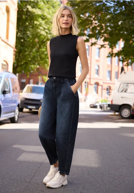 Eine Frau steht auf einer sonnigen Straße vor parkenden Autos, trägt ein schwarzes Top, weite Jeans und weiße Sneaker.