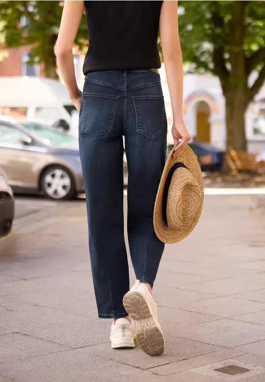 Person in Jeans, schwarzem Top und weißen Sneakern, läuft den Bürgersteig entlang und hält einen Strohhut.