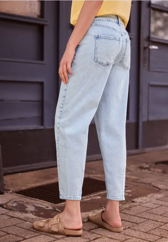 Rückenansicht einer Person in hellblauen Jeans und flachen Sandalen auf gepflastertem Gehweg.