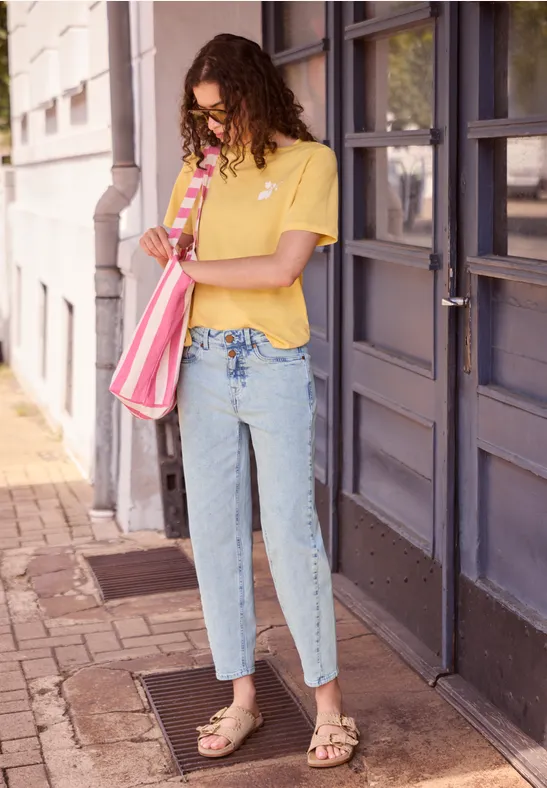 Frau mit gelbem T-Shirt, blauen Jeans und Sandalen steht mit rosafarbener Tasche vor einer Haustür.
