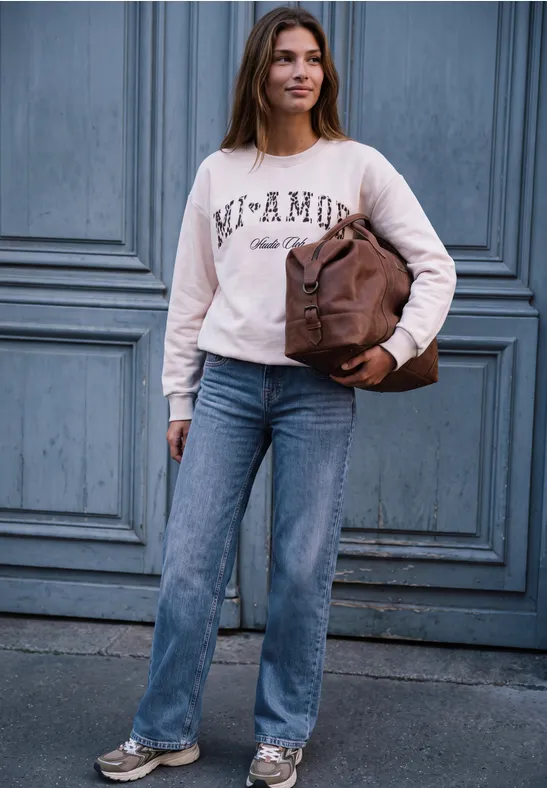 Junge Frau steht vor blauer Holztür, trägt Jeans, ein Sweatshirt mit Aufschrift und hält eine braune Tasche.