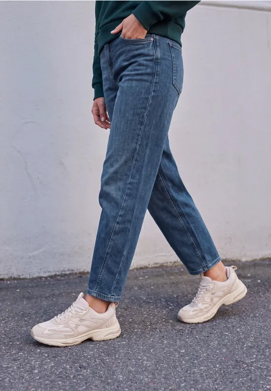 Persoon in blauwe jeans en witte sneakers loopt langs een muur.