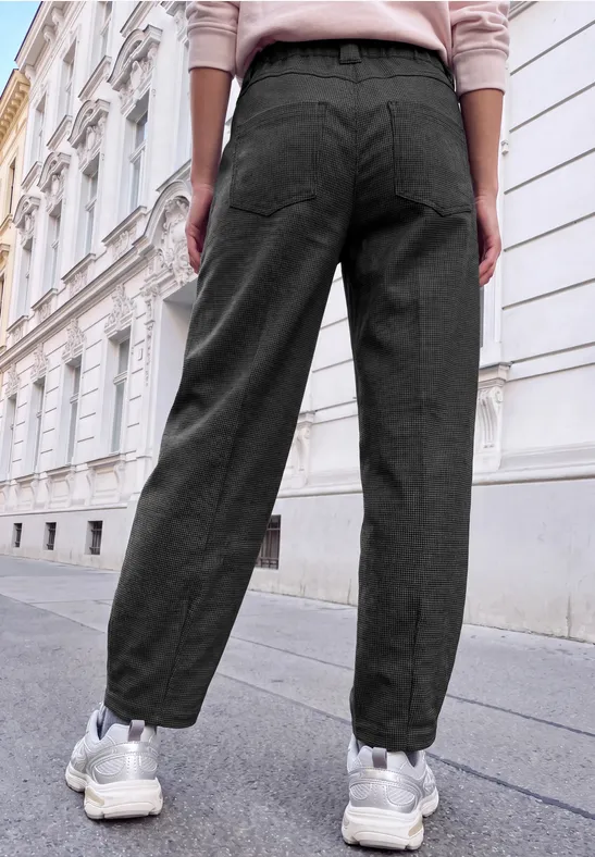 Eine Person steht in schwarzen Stoffhosen und weißen Sneakern vor einer historischen Hausfassade.