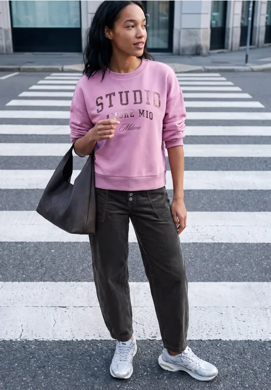 Jeune femme portant un sweat rose et un sac en bandoulière, debout sur un passage piéton en ville.