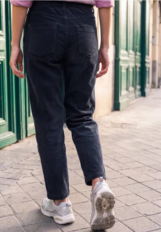 Person in schwarzen Jeans und weißen Sneakers von hinten auf einem Gehweg vor grünen Türen.