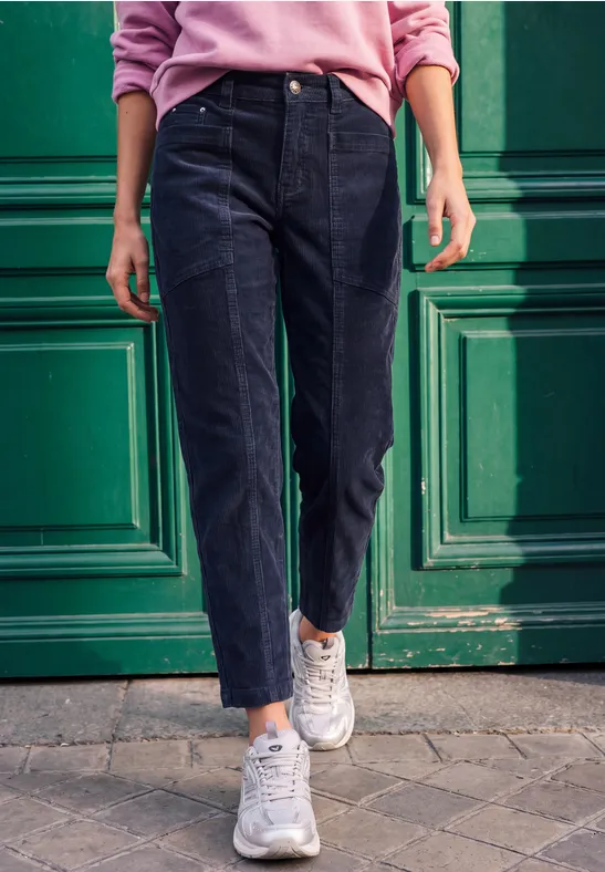 Person im rosa Pullover, schwarzer Jeans und weißen Sneakern steht vor einer grünen Tür.