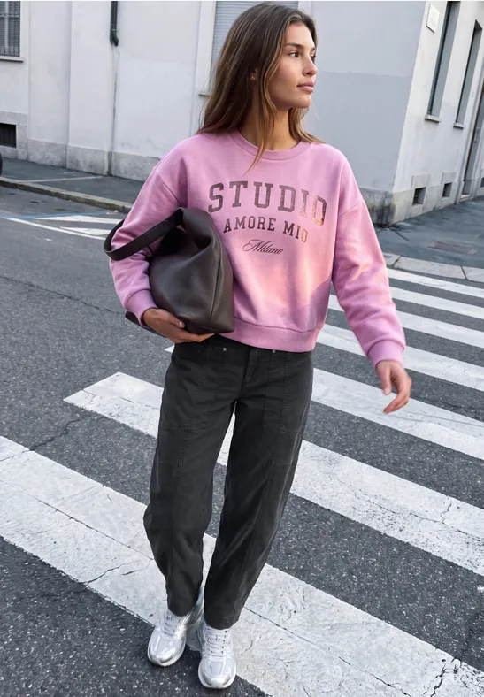 Frau mit rosa Sweatshirt, dunkelgrauen Hosen und Turnschuhen überquert Zebrastreifen, trägt Tasche.