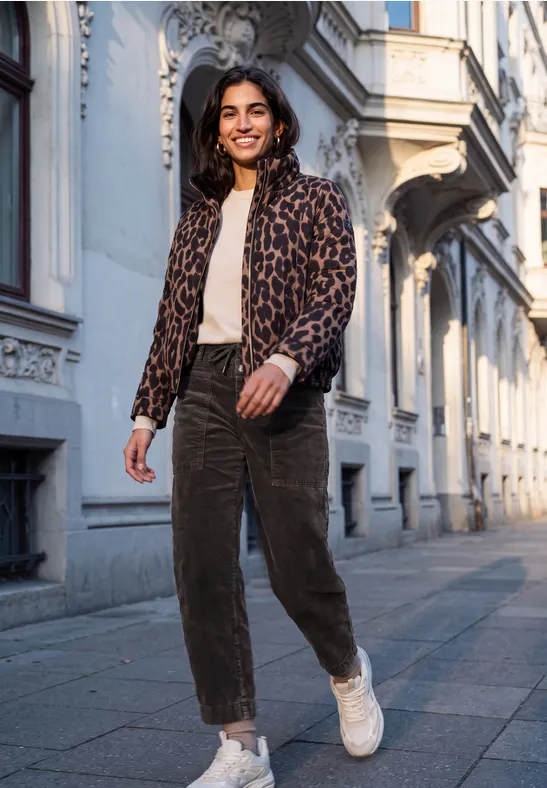 Eine junge Frau in Leo-Jacke und weißen Sneakern geht lächelnd eine Straße mit historischer Architektur entlang.