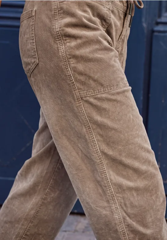 Braune Cordhose an Personenbeinen, seitliche Nahaufnahme vor dunklem Hintergrund.