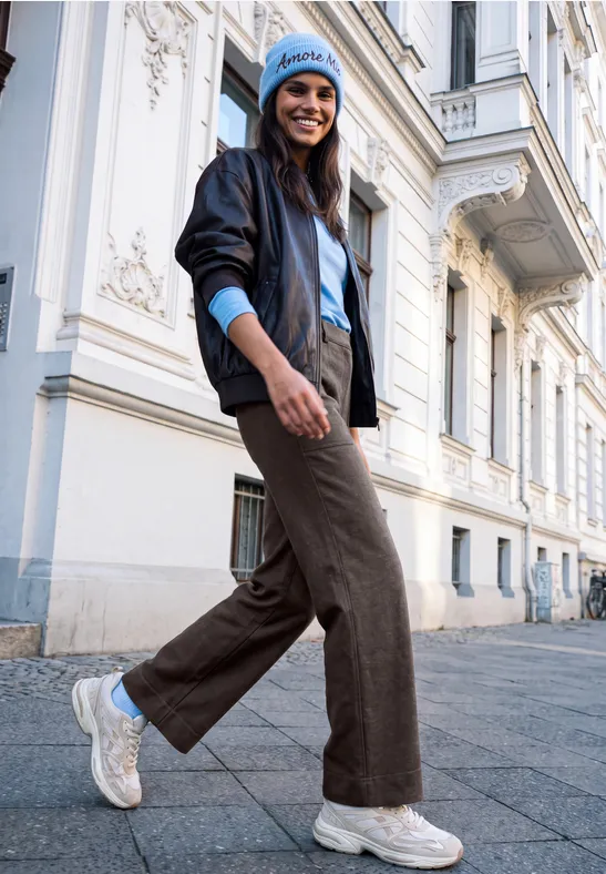 Junge Frau in stylischer Jacke, Mütze und breiten Hosen spaziert lächelnd vor historischer Fassade.