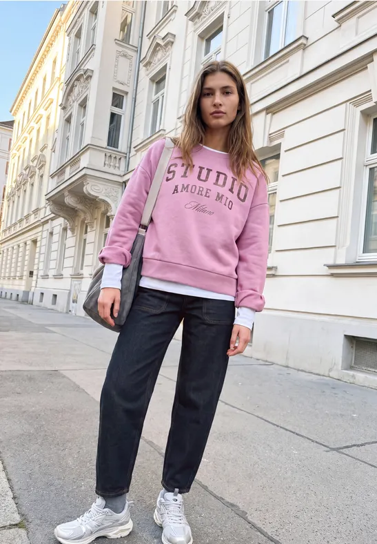 Junge Frau in rosa Sweatshirt, schwarzer Hose und weißen Sneakern steht auf Gehsteig vor Altbau.
