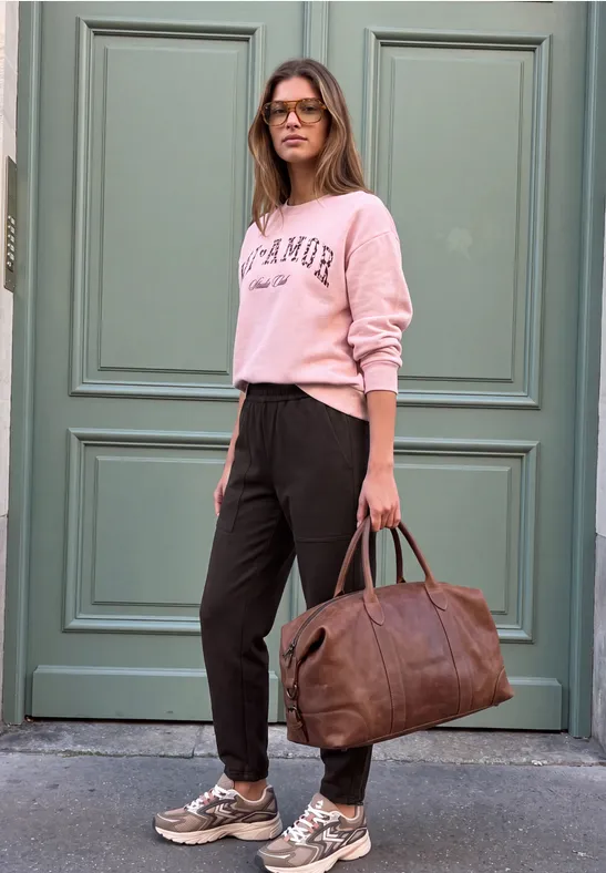 Frau in lässigem Outfit mit rosa Pullover und schwarzer Hose, trägt eine braune Reisetasche vor einer grünen Tür.