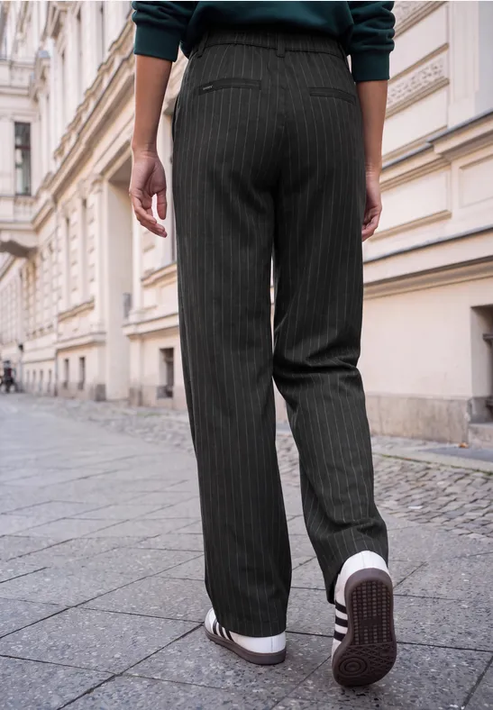 Person in dunklen Nadelstreifenhosen und weißen Sneakers geht auf einem Gehweg an historischen Gebäuden entlang.