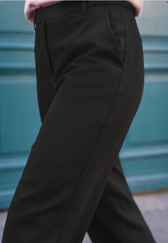 Nahaufnahme von schwarzen Stoffhosen mit Bügelfalte vor türkisblauem Hintergrund.