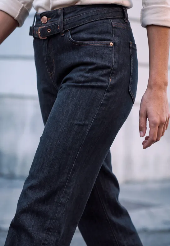 Vue partielle d’une personne en jean bleu foncé avec ceinture, debout devant un arrière-plan flou.