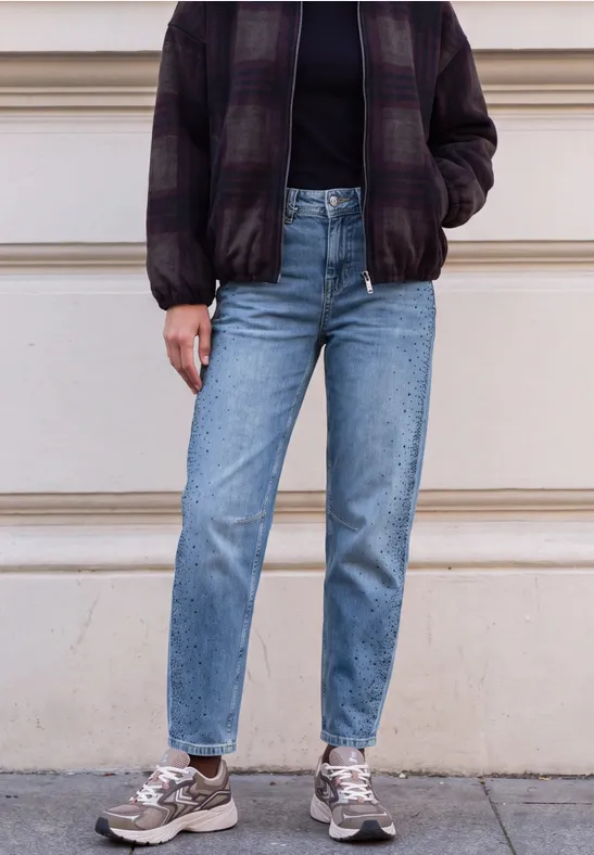 Persoon met paarse geruite bomber, zwarte trui, blauwe jeans en grijze sneakers voor een gevel.