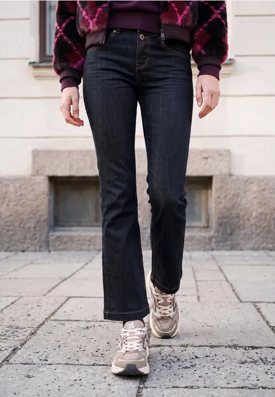 Persoon in zwarte jeans met licht uitlopende pijpen, grijs-beige sneakers en een gebreid jasje.