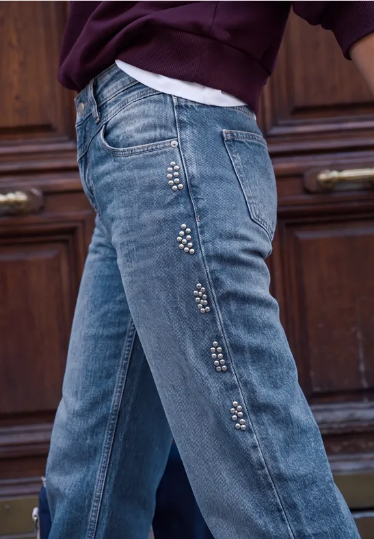 Hüfte einer Person in blauen Jeans mit Nieten entlang der Seitennaht, Hintergrund Holztür.