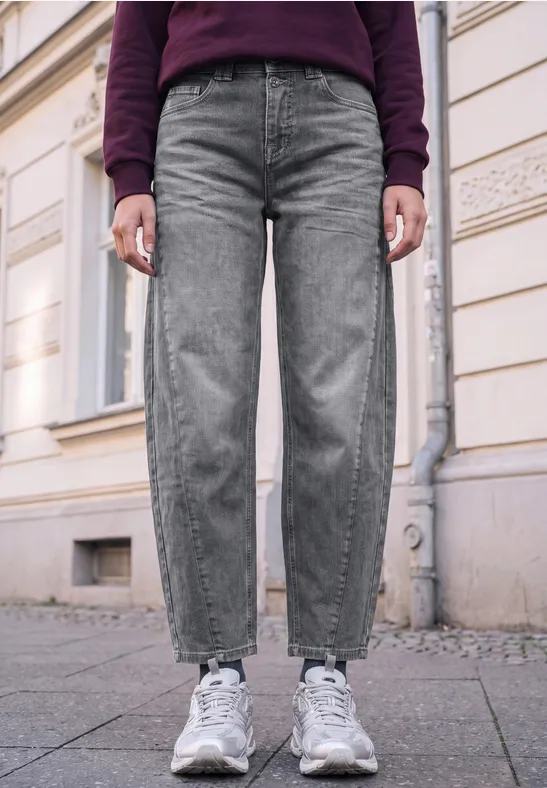 Unterkörper einer Person in grauen Jeans und weißen Sneakern auf Bürgersteig vor einer Hauswand.