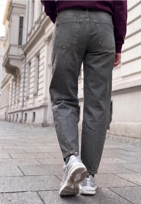 Eine Person in grauen Jeans und weißen Sneakers geht auf Kopfsteinpflaster an einem alten Gebäude entlang.