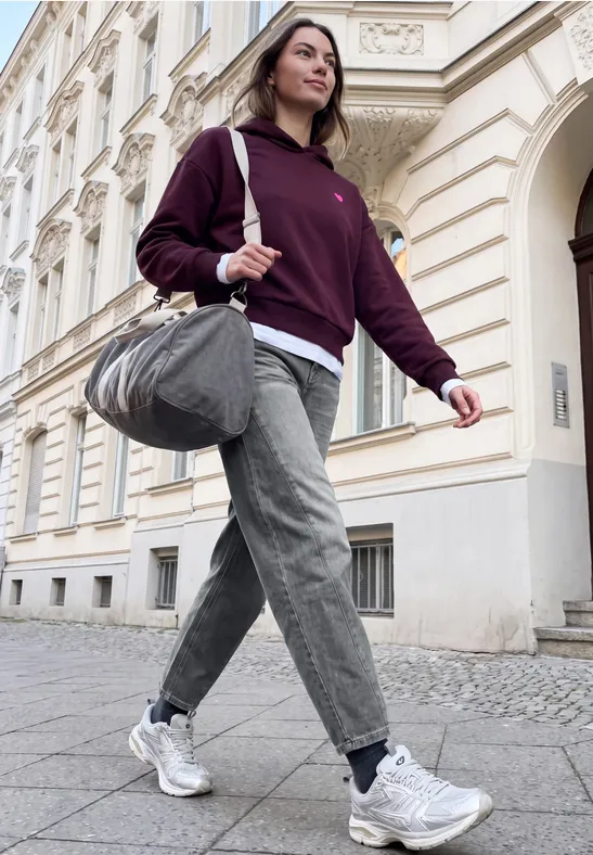 Eine junge Frau in Kapuzenpullover, Jeans und Sneakern trägt eine Sporttasche und geht eine Straße entlang.