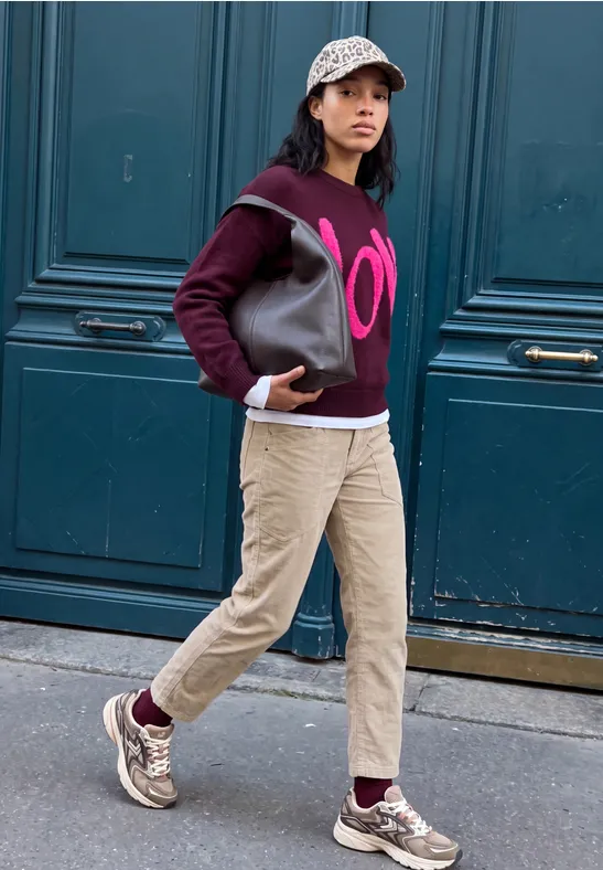 Eine junge Frau in Hoodie, Cargohose und Sneakern geht mit Tasche an einer blauen Tür vorbei.