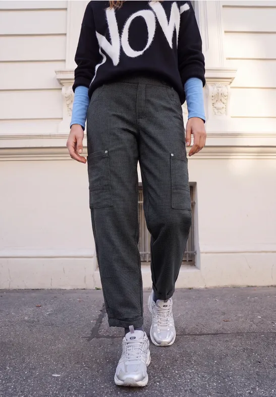 Personne en pantalon cargo kaki, pull noir 'NOW', sous-pull bleu et baskets blanches, marchant sur un trottoir.