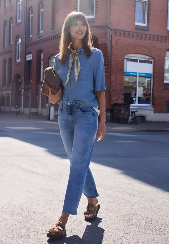 Une femme en jean, chemise et sandales marche en ville avec un sac à bandoulière marron.