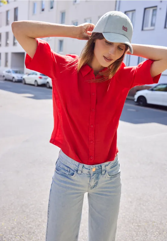 Person mit rotem Hemd und Jeans, trägt Baseballkappe, steht auf sonniger Straße und hält Hände hinter Kopf.