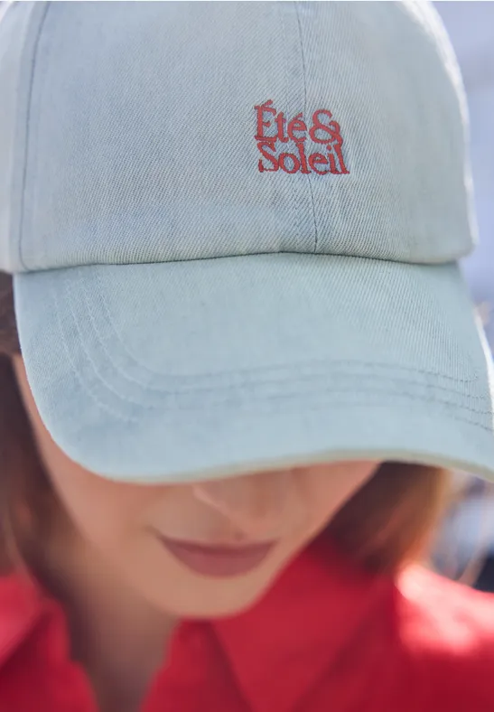 Eine Person trägt ein hellblaues Jeanscap mit roter Aufschrift 'Été & Soleil'. Gesicht unscharf.